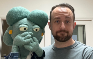 Jason Fluegge's headshot, Jason is smiling while holding a green puppet with it's hands over it's mouth, possibly laughing